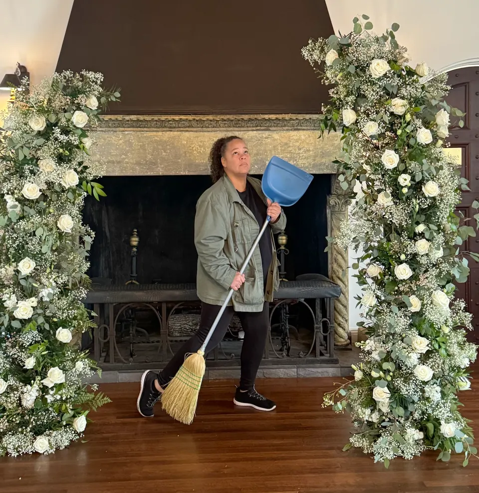 Jackie, our Event Lead, playfully posing with a broom between two white rose floral columns