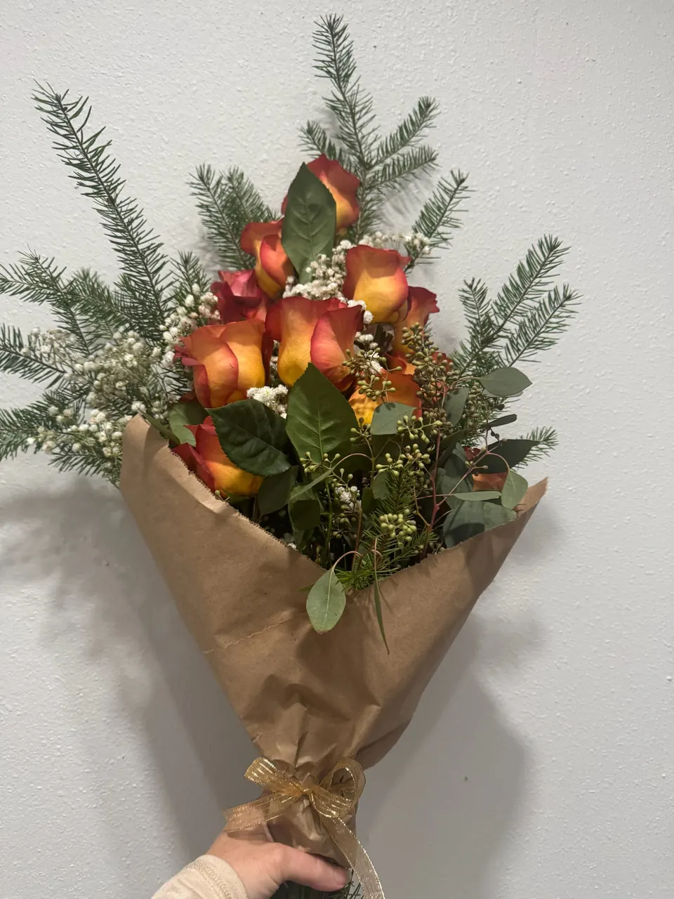 A kraft-wrapped bouquet of orange-red roses with evergreen sprigs, baby’s breath and a gold ribbon
