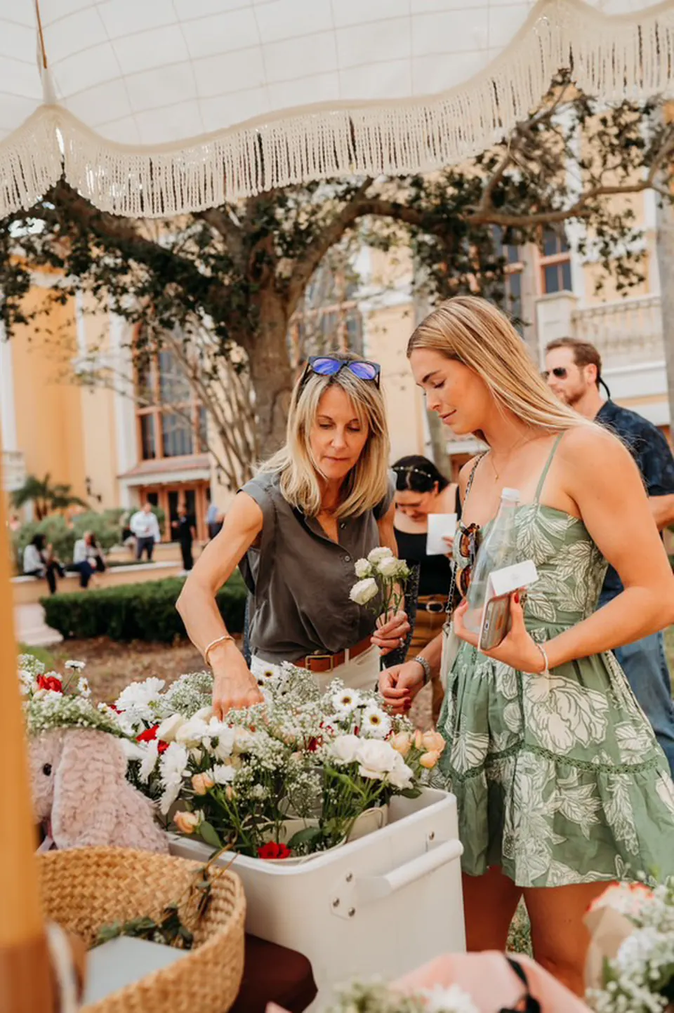 Two guests choosing white daisies and fresh blooms from a flower cart under a fringed umbrella